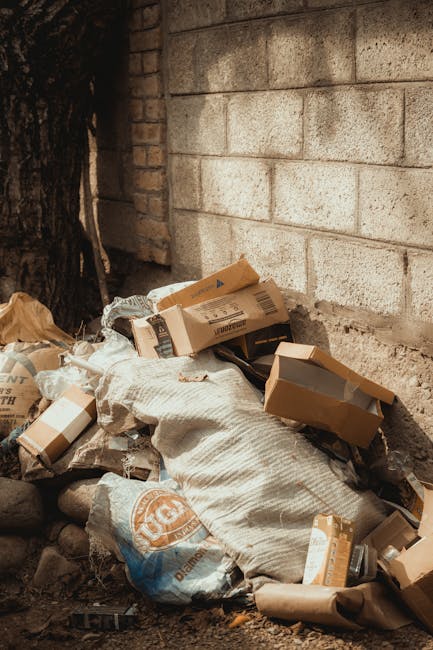 A pile of waste materials sits against a brick and concrete wall. The collection includes flattened cardboard boxes, some with printed labels and barcodes, a large white sack made of textured fabric, and various bits of brown paper and packaging debris. The cardboard boxes are mostly brown with some partially open and leaning on each other; the sack appears to be filled with additional rubbish. The environment suggests an outdoor area, possibly a back alley or service yard, with the waste accumulated near the base of a weathered wall. There is a large dark tree trunk or post on the left side, partially obscuring the waste. The scene features natural daylight illuminating the materials, highlighting textures like the corrugated surfaces of the boxes and the coarse fabric of the sack. This image illustrates a scenario of private or independent waste collection outside a building, aligning with rubbish removal or on-site clearance services provided by Waste Removal Wandsworth.