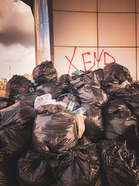 Near the edge of a paved street, three large black plastic trash bags are stacked against a black metal fence with vertical bars, which extends into the background. The bags appear to be filled with rubbish, showing creases and wrinkle textures on their surfaces. To the left of the bags, a bush with dense, leafy foliage partially obscures the scene, while a tree with dark leaves rises above the fence. The environment is an outdoor urban setting, with the bags placed on the curb in front of the fence, possibly indicative of private or alternative waste disposal methods. The lighting is dim, suggesting early morning or late evening, with soft shadows cast across the scene. The overall impression indicates an instance of rubbish waiting for collection, typical of independent waste removal services, as it aligns with contextually relevant waste management practices.