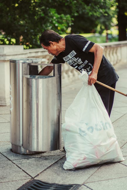 A middle-aged man with dark hair is standing outdoors on a paved walkway, leaning forward and placing a large white plastic bag filled with rubbish into a stainless steel public rubbish bin. The bag appears to contain debris or waste material and is partially transparent, with some visible contents inside. The man's posture suggests he is carefully depositing the waste, and he is dressed in a black T-shirt with white printed text on the front and dark trousers. In his right hand, he holds a wooden stick or broom handle, while his left hand guides the bag into the bin. Behind him, there is a low stone or concrete balustrade, and lush green trees and bushes provide a natural backdrop, indicating a park or urban outdoor setting. The lighting is natural and evenly distributed, highlighting the metallic finish of the rubbish bin and the details of the pedestrian walkway. The scene exemplifies routine street waste disposal, which can be part of an alternative waste handling service overview, as provided by Waste Removal Wandsworth, supporting the context of private rubbish collection and on-site waste clearance in urban environments.