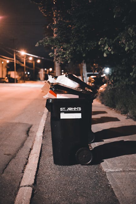 A black wheeled rubbish bin positioned on the sidewalk beside a curb in an outdoor urban setting at night, with a white label on the front that reads 'ST. JOHN'S' and partially visible text beneath it. The bin is open at the top, with various types of waste, including paper and cardboard, resting inside and leaning against the rim. The surrounding environment includes a street lightly illuminated by streetlights, casting warm light and shadows on the pavement and the bin. In the background, there are trees, parked vehicles, and additional streetlights, suggesting a residential or mixed-use area. The scene reflects a typical private waste collection scenario, where bins are left near the curb for collection, indicative of independent rubbish disposal services. The details in the image highlight the texture of the waste materials, the smooth finish of the bin, and the nighttime atmosphere of the collection process.