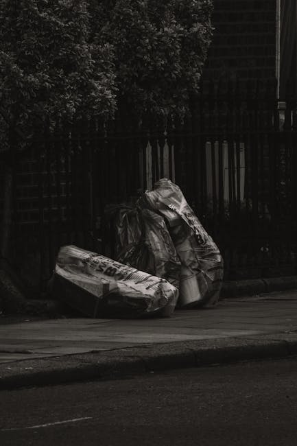 A middle-aged man with dark hair is standing outdoors on a paved walkway, leaning forward and placing a large white plastic bag filled with rubbish into a stainless steel public rubbish bin. The bag appears to contain debris or waste material and is partially transparent, with some visible contents inside. The man's posture suggests he is carefully depositing the waste, and he is dressed in a black T-shirt with white printed text on the front and dark trousers. In his right hand, he holds a wooden stick or broom handle, while his left hand guides the bag into the bin. Behind him, there is a low stone or concrete balustrade, and lush green trees and bushes provide a natural backdrop, indicating a park or urban outdoor setting. The lighting is natural and evenly distributed, highlighting the metallic finish of the rubbish bin and the details of the pedestrian walkway. The scene exemplifies routine street waste disposal, which can be part of an alternative waste handling service overview, as provided by Waste Removal Wandsworth, supporting the context of private rubbish collection and on-site waste clearance in urban environments.